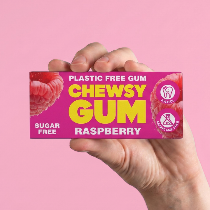 A hand holding a pink box of Chewsy Gum Raspberry flavor against a solid pink background.