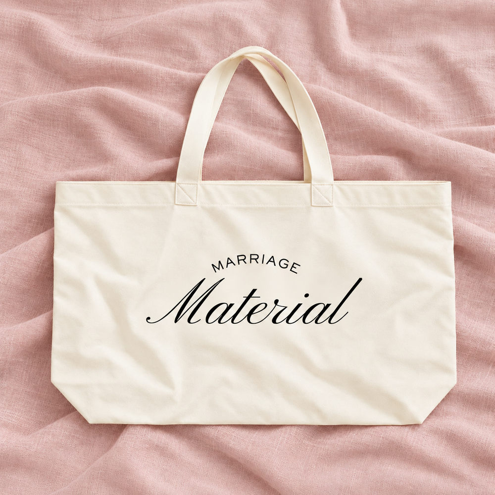 An off-white canvas tote bag with black printed text reading 'MARRIAGE Material' on a soft pink fabric background.