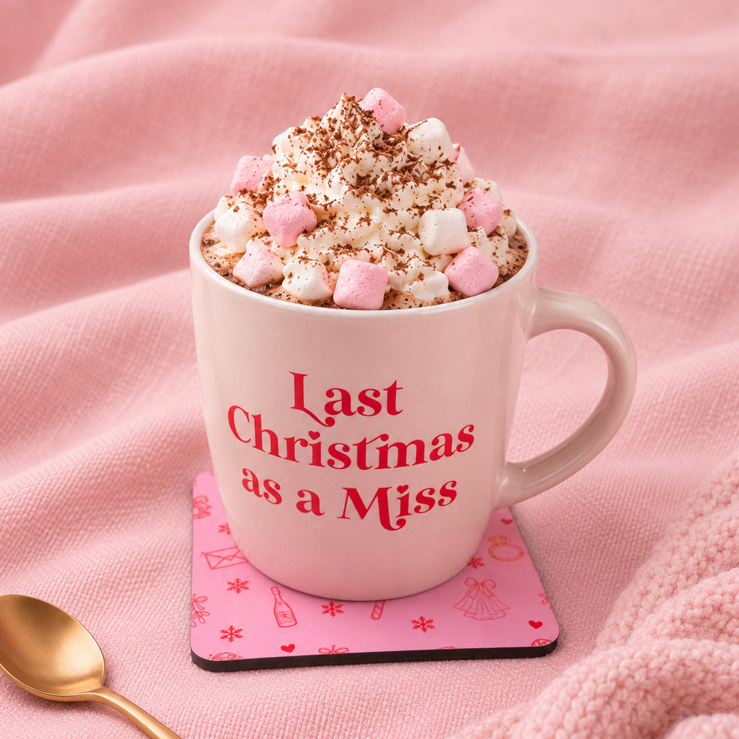A pink ceramic mug with the phrase 'Last Christmas as a Miss' in red text, filled with whipped cream, marshmallows, and cocoa powder, set on a pink coaster with a gold spoon on a pink textured blanket.