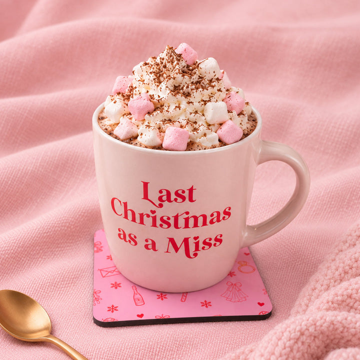 A pink ceramic mug with the phrase 'Last Christmas as a Miss' in red text, filled with whipped cream, marshmallows, and cocoa powder, set on a pink coaster with a gold spoon on a pink textured blanket.