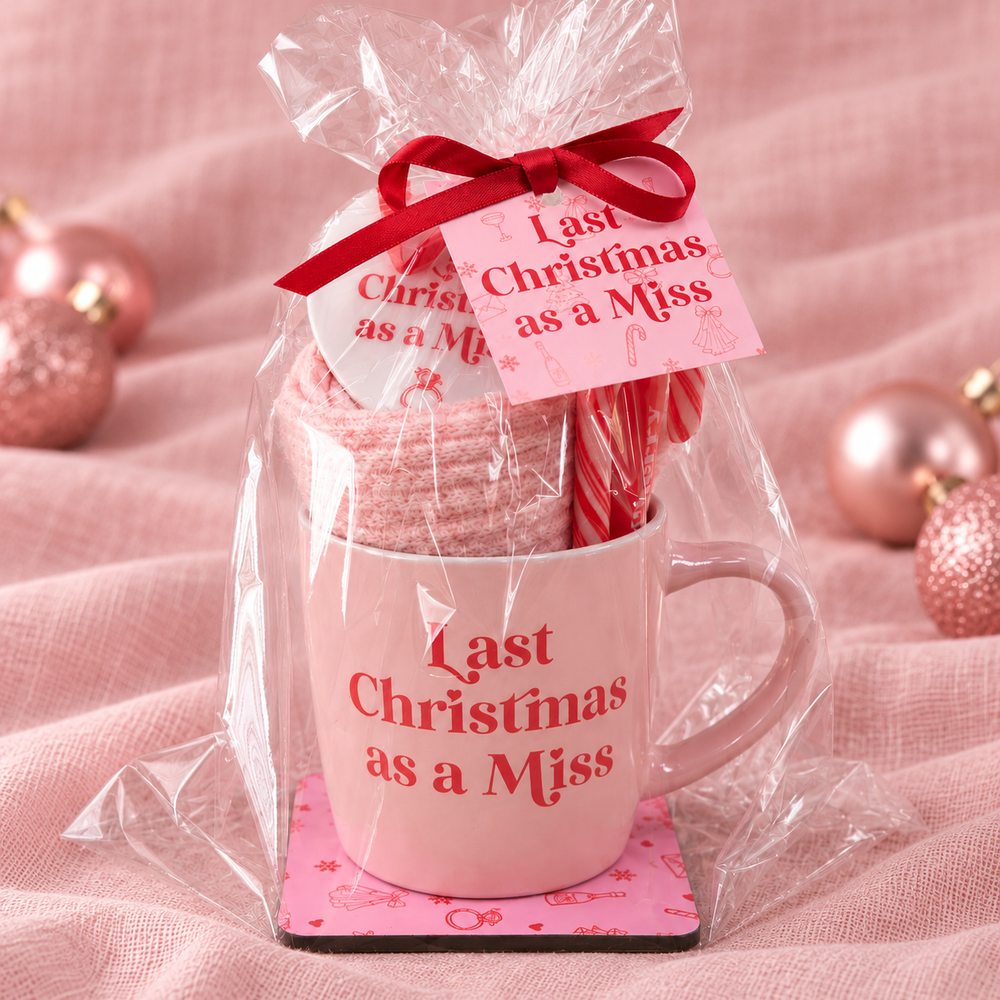 A Christmas-themed gift set in clear cellophane packaging, featuring a light pink ceramic mug with red text reading 'Last Christmas as a Miss,' a pink gift tag with matching text, a red ribbon bow, a pink striped candy cane, and a folded pink knit item, arranged on a pink fabric backdrop with decorative rose-gold ornaments.