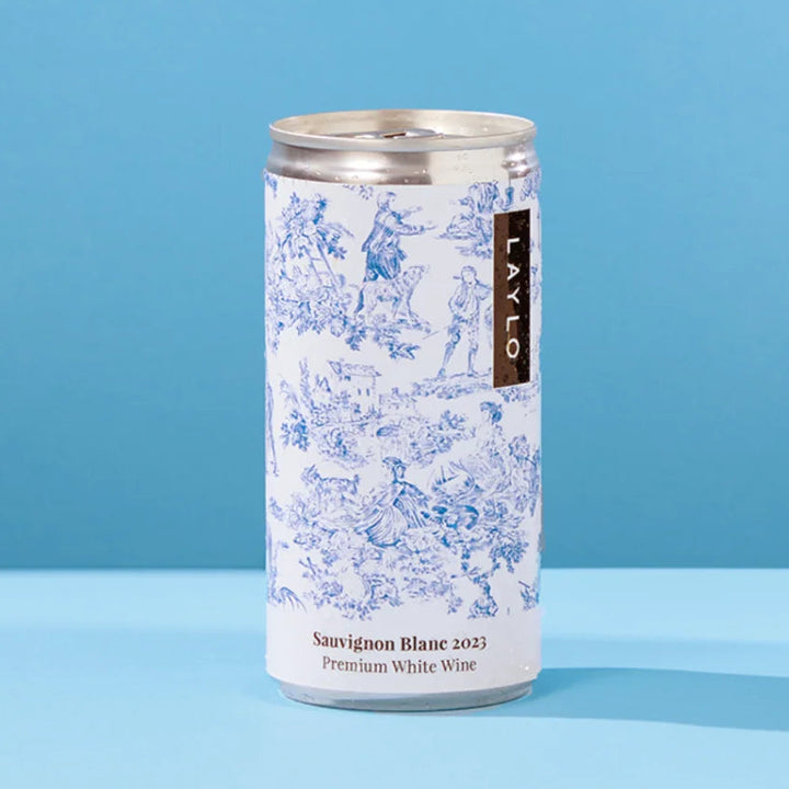 A single can of Laylo Silver Edron Rhine 2023 Premium White Wine stands on a light blue surface against a solid blue background. The can features a white label with a blue floral pattern and a vertical black brand label.