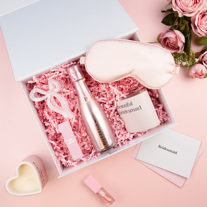 A bridal shower gift set arranged in an open white box with pink shredded paper. The set includes a pale pink satin sleep mask, a metallic rose-gold bottle, a small pink candle, a lip gloss tube, and cards.
