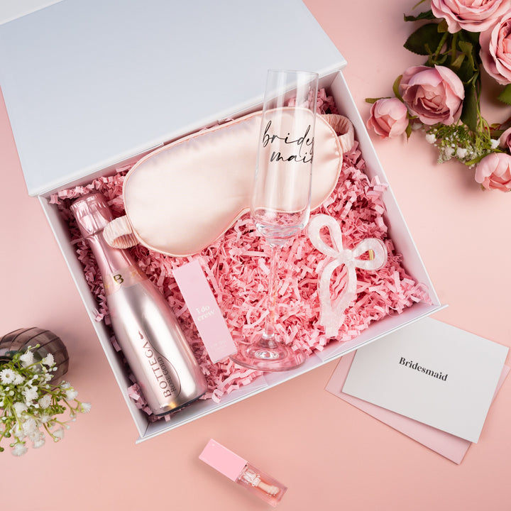 Bridesmaid gift box set on a pink background, containing a champagne bottle, clear champagne flute, satin sleep mask, and lip gloss, with a card reading 'Bridesmaid'.