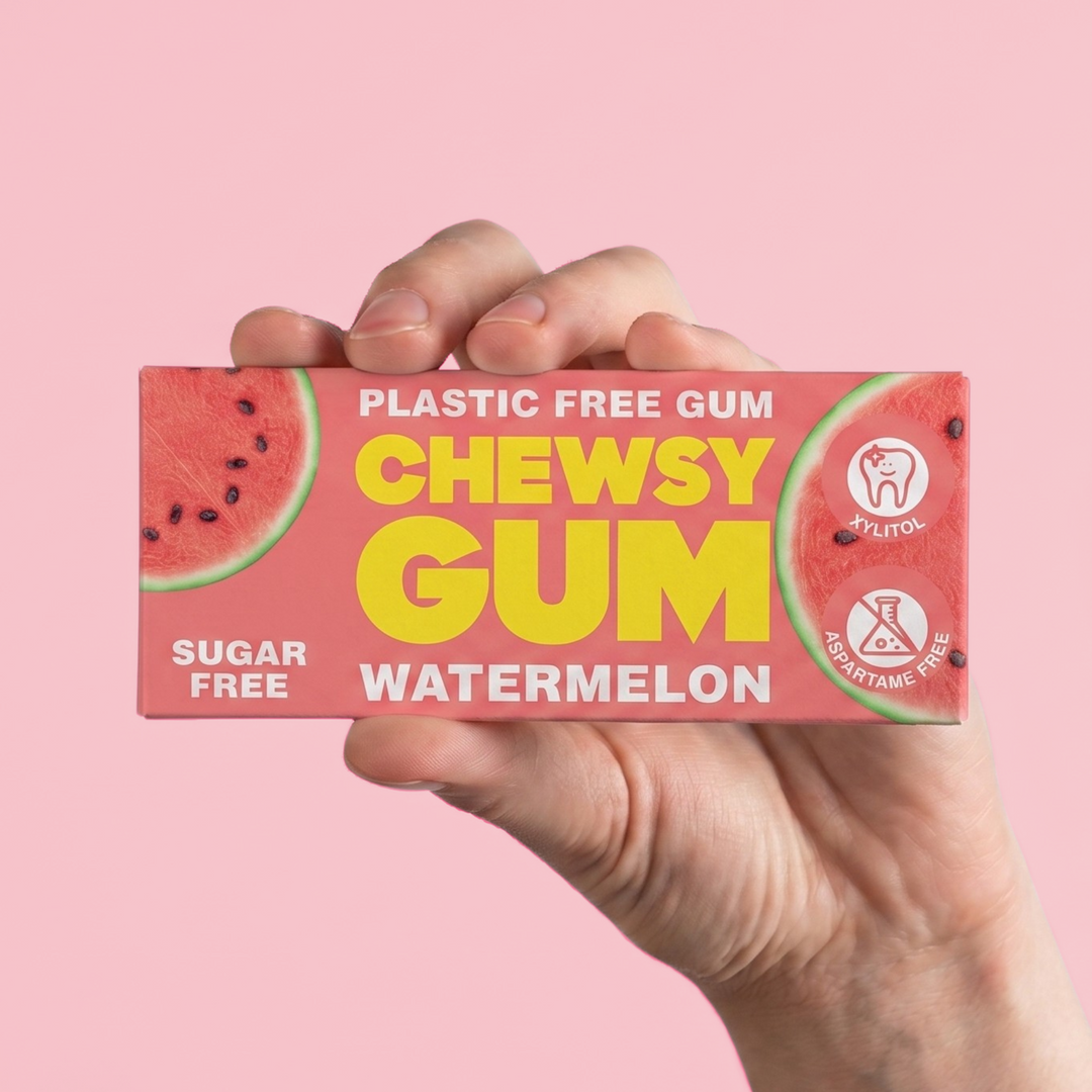 A hand holding a rectangular box of watermelon-flavored chewy gum against a solid pink background. The box features a watermelon graphic and bold yellow text reading 'CHEWSY GUM' with 'WATERMELON' underneath.