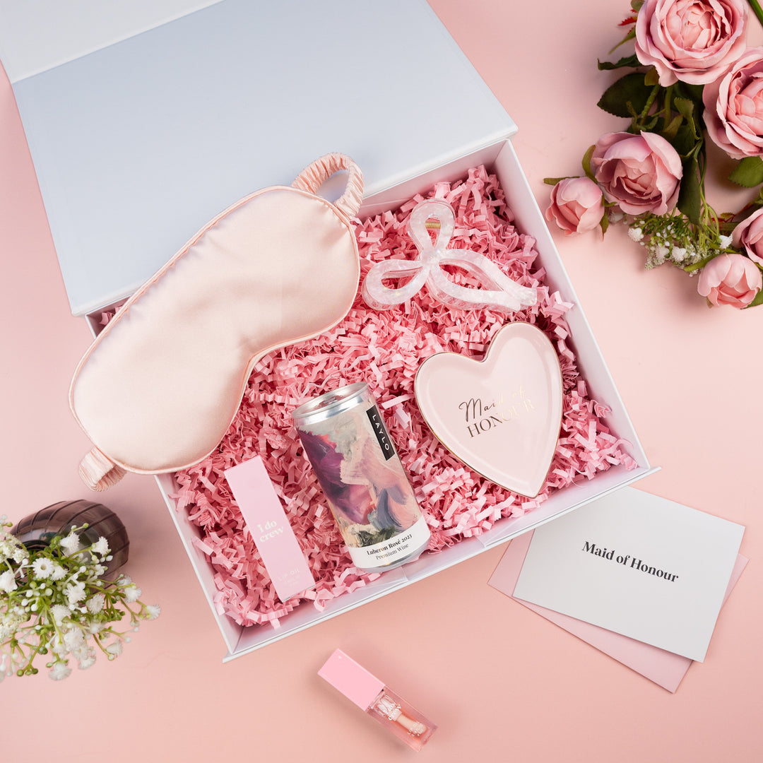 A styled flat-lay gift box set on a soft pink background, featuring a pale pink satin sleep mask, a heart-shaped soap, a small can, a pink lip product, and a white card reading 'Maid of Honour' nestled in pink crinkle paper inside an open white box.